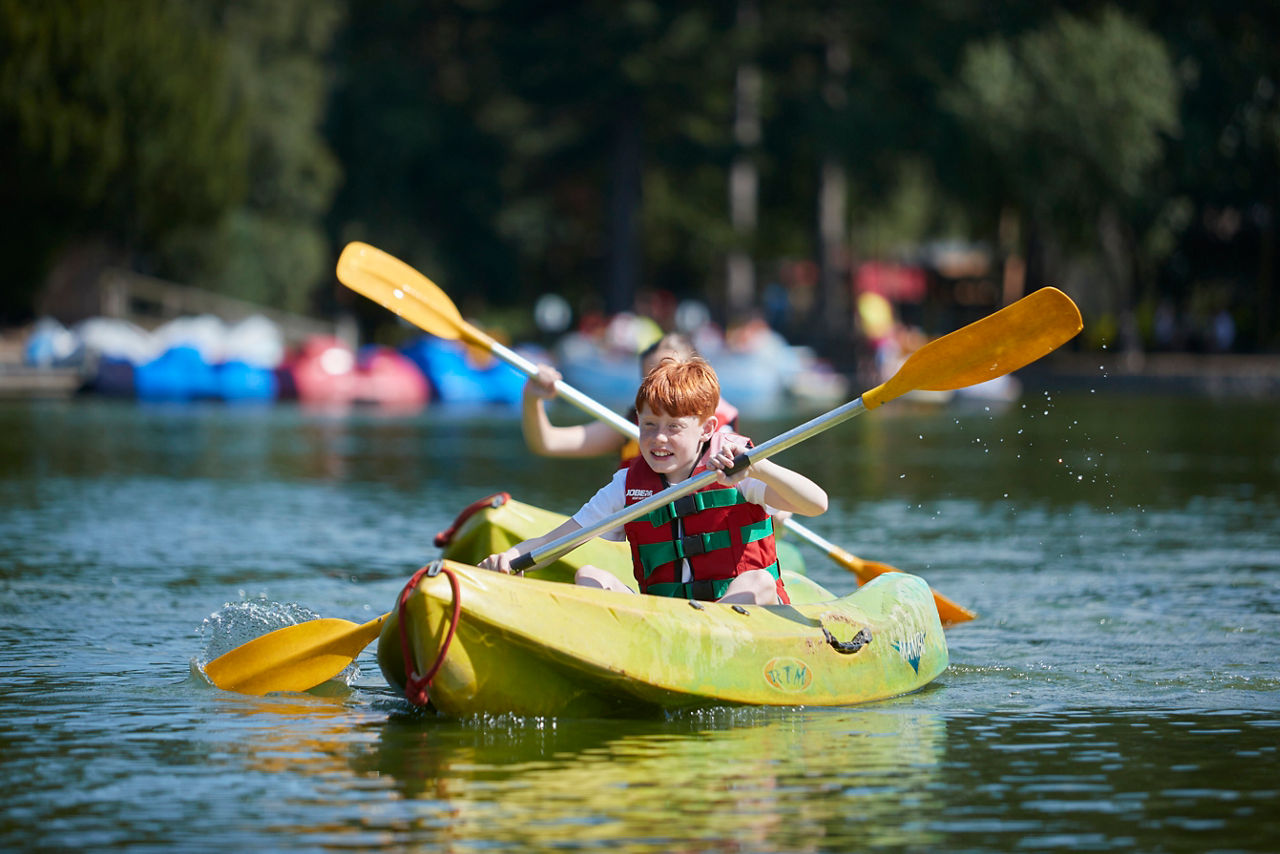 Two children in a yellow kayak paddle forward on a calm lake, wearing life vests; splashes trail their oars; distant boats and trees blur behind. Visible text: RTM.